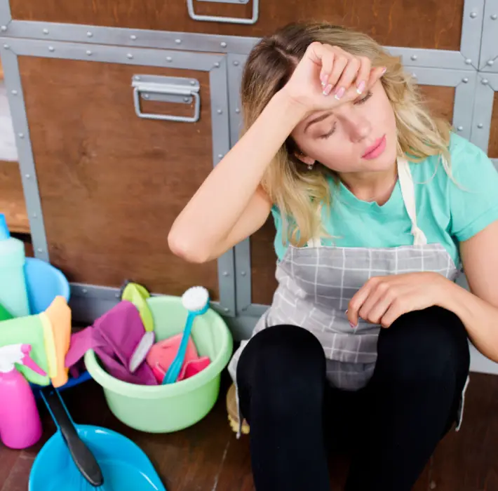 Exhausted woman in apron resting after DIY bond cleaning with supplies scattered around — illustrating the stress of end-of-lease cleaning without professional help